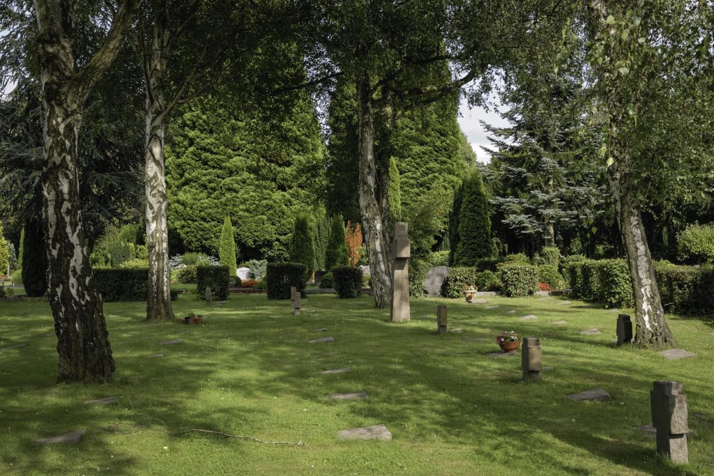 Ein friedlicher Friedhof mit verstreuten Grabsteinen, umgeben von hohen Bäumen und üppigem grünen Gras, unter einem teilweise bewölkten Himmel. Blumen und Sträucher bringen Farbe in die ruhige Landschaft.